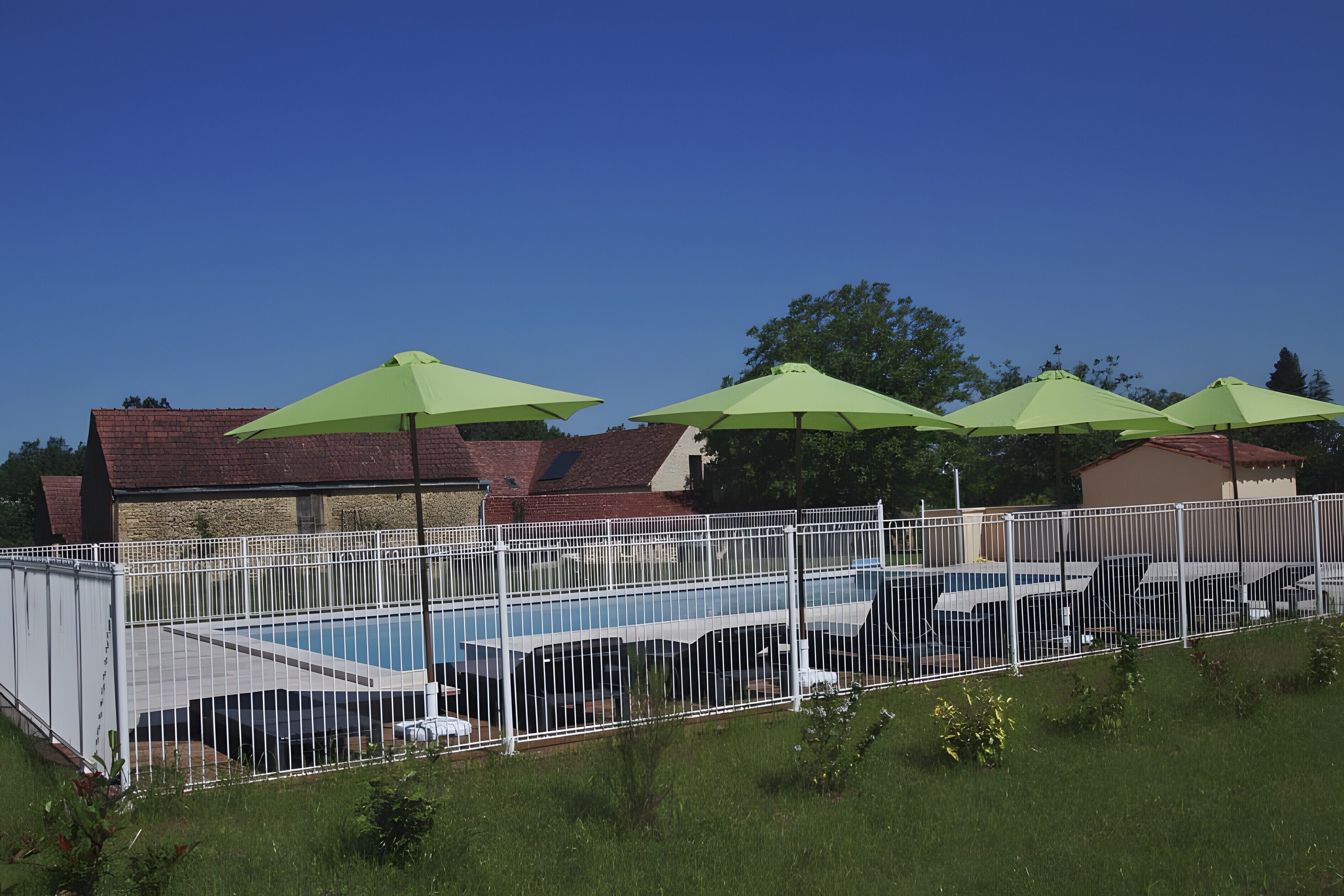 Ein Schwimmbecken mit grünen Sonnenschirmen, umgeben von einem weißen Zaun, mit Gebäuden im Hintergrund.