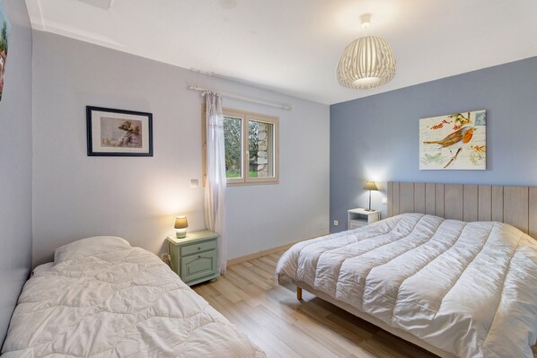 Une chambre avec deux lits, une fenêtre avec des rideaux, une table de chevet avec une lampe et un mur orné d’un tableau encadré.