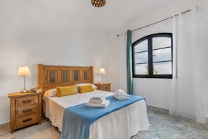 Une chambre à coucher avec une tête de lit en bois, deux tables de chevet avec des lampes, un lit recouvert d’une couvre-lit bleue et blanche, et une fenêtre avec des rideaux.
