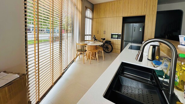 A modern kitchen with wooden cabinets, a built-in microwave, and a stainless steel refrigerator. There is a bicycle parked near the window, and a table with chairs is visible in the background.
