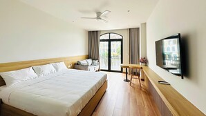 A hotel room with a large bed, a desk with a chair, a television, and a balcony with curtains.