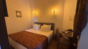 Una cama individual con una colcha estampada, dos lámparas de pared, una mesita con una silla y un cuadro en la pared.