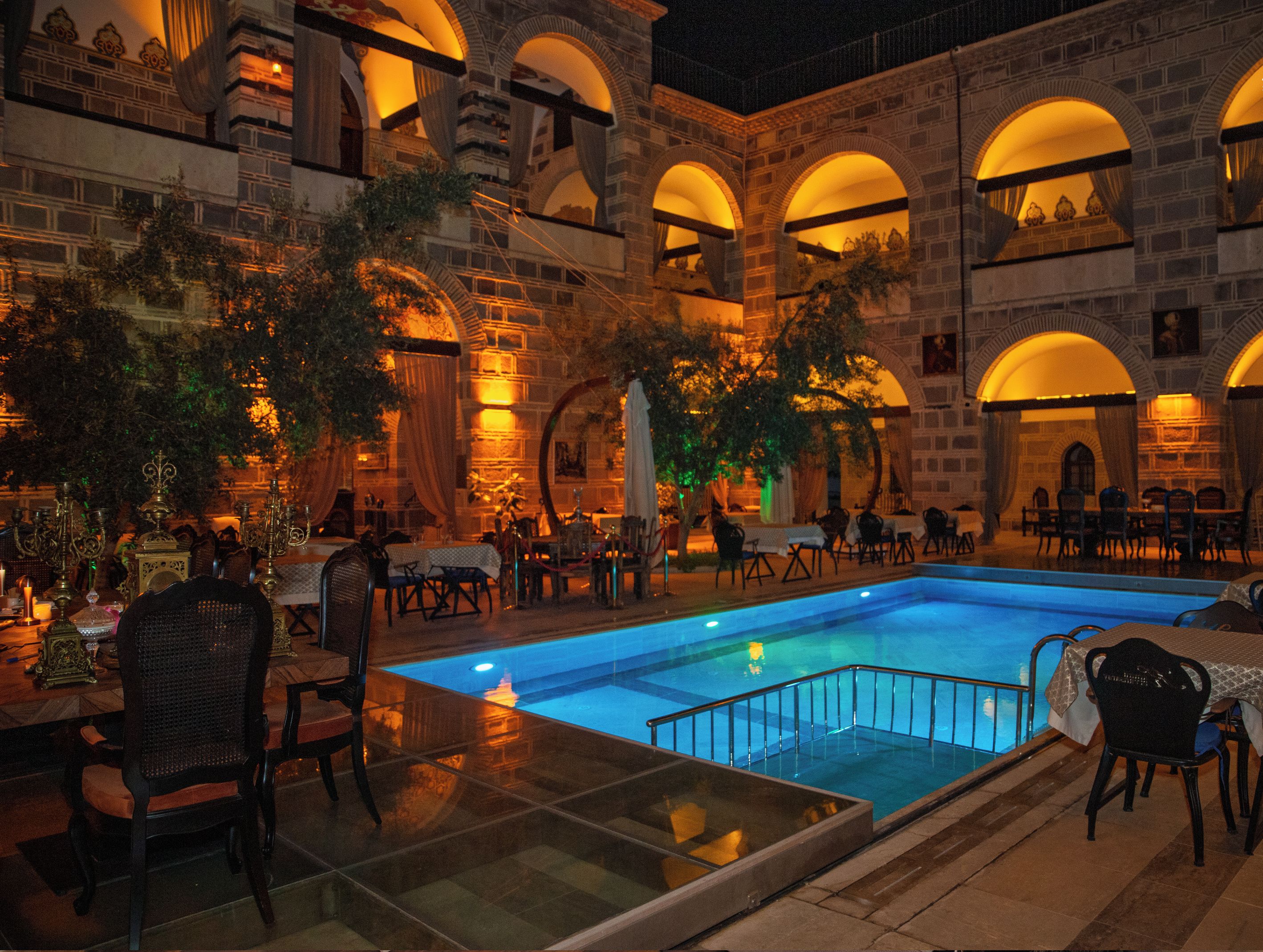 Un área junto a la piscina con mesas y sillas, una piscina y un edificio con arcos y un letrero que dice "KERVANSARAY".