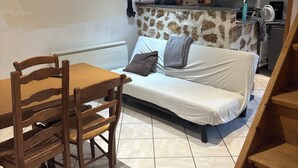 A cozy room with a wooden dining table and chairs, a sofa with a stone accent wall, and a kitchen area with a fan and various items on the counter. - Apparently from the countryside (La Croix-sur-Ourcq)