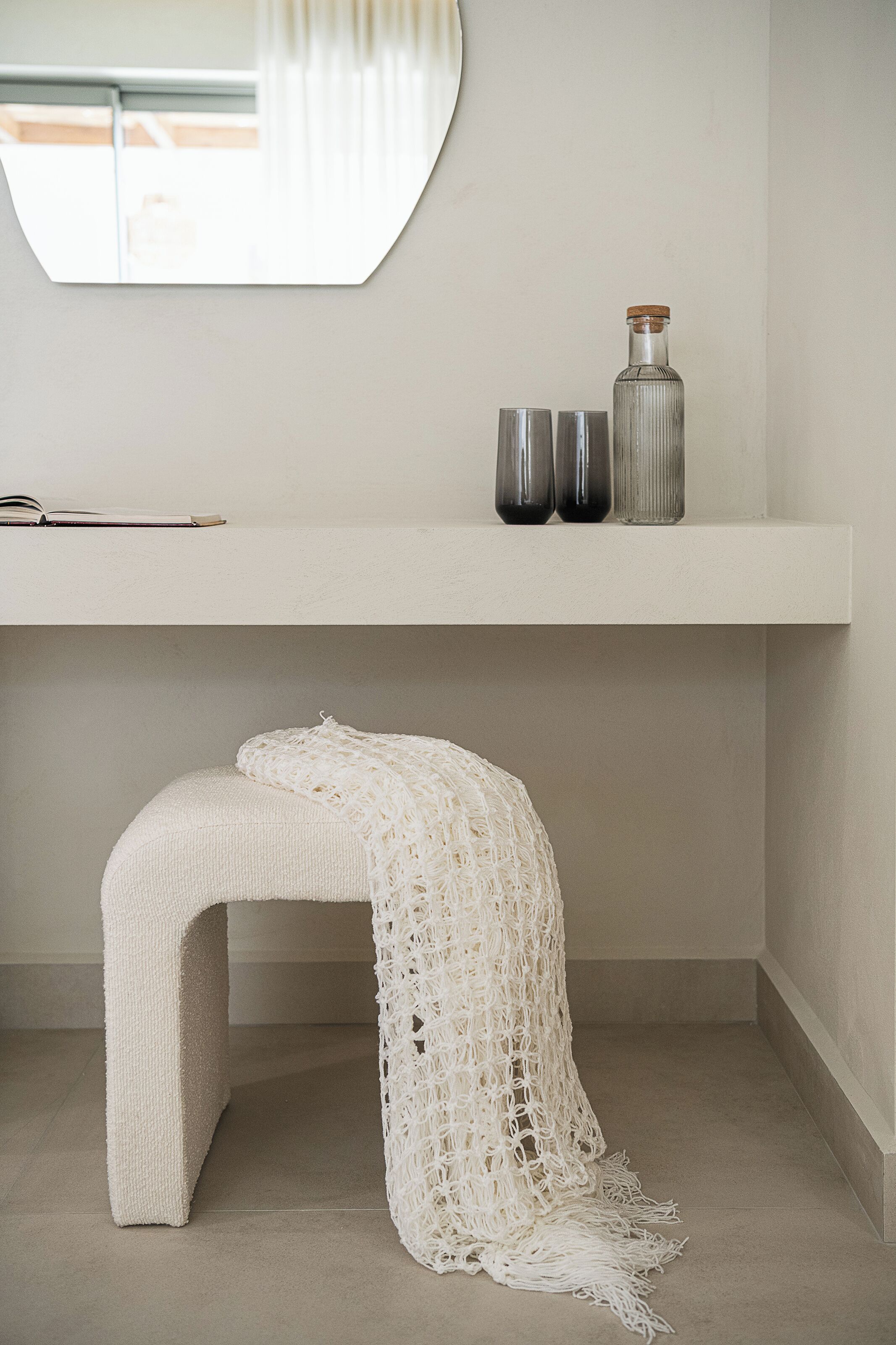 Une salle de bain minimaliste avec un banc blanc, un miroir et une étagère sur laquelle se trouvent une bouteille et deux verres.
