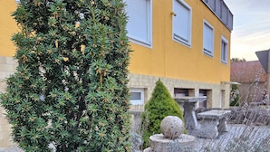 Ein gelbes Gebäude mit weißen Fensterläden, einer Steinfontäne und einer Nadelbaumart.