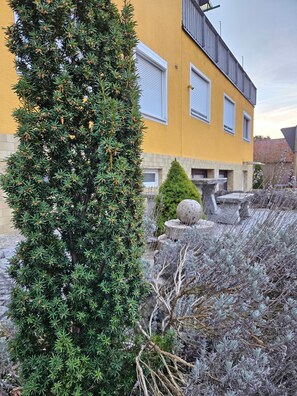 Un bâtiment jaune avec des volets blancs, une fontaine en pierre et un arbre à feuilles persistantes.