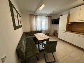 Un espace de vie compact comprenant un coin cuisine, un espace repas avec une banquette, un téléviseur fixé au mur et une fenêtre avec des rideaux.