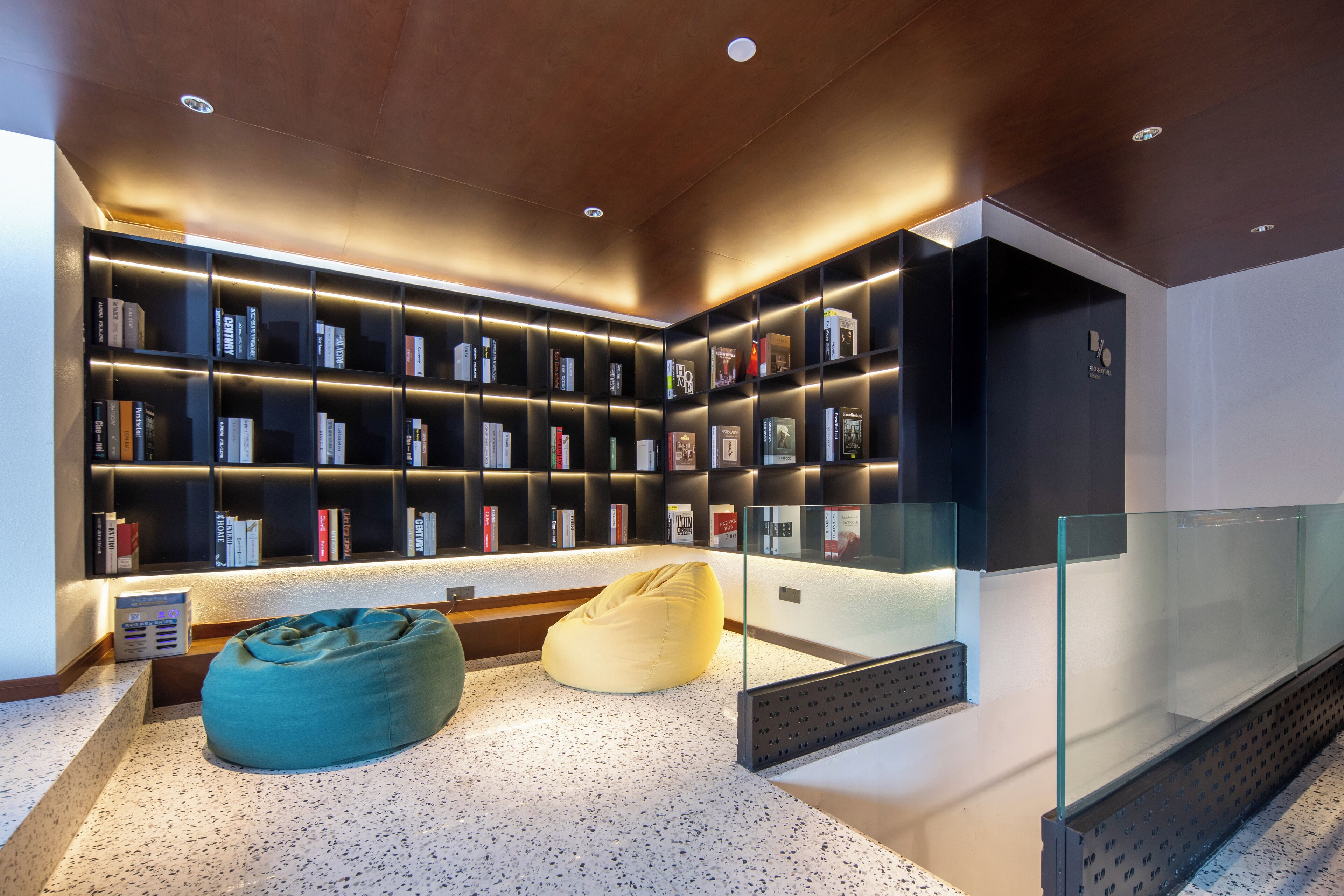 Un área de lectura moderna con estanterías integradas, barandilla de vidrio y dos pufs.