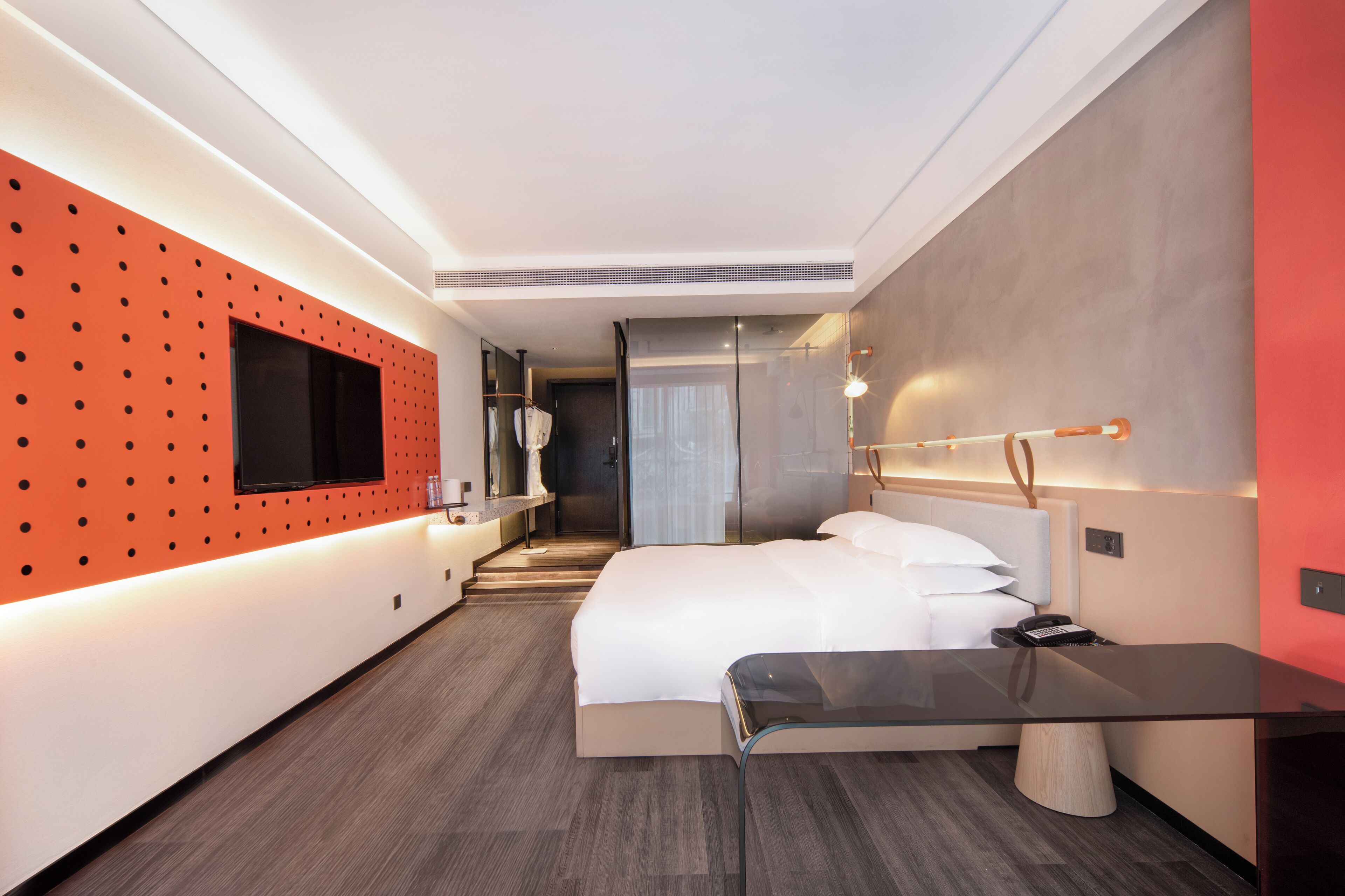A modern hotel room with a large bed, a glass desk, a red accent wall, and a TV mounted on the wall.