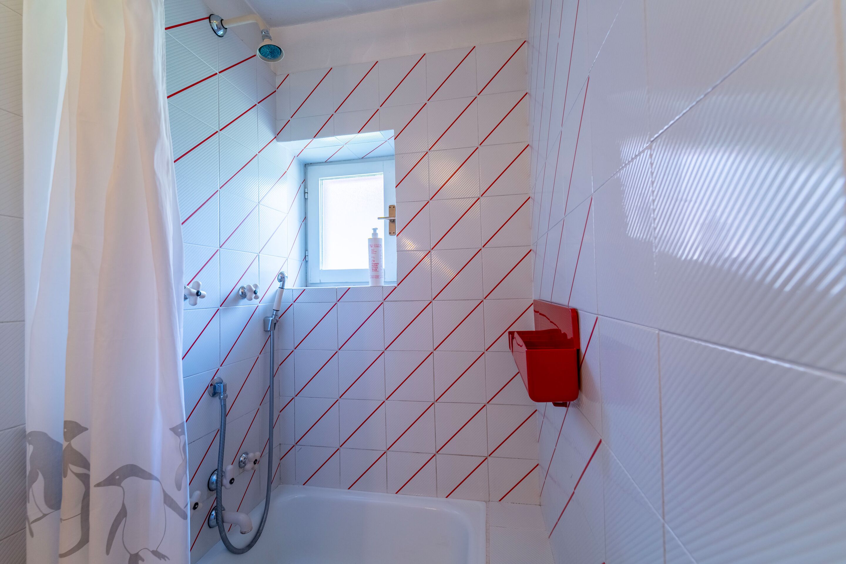 Un baño con paredes de azulejos blancos, una ducha, una ventana y un perchero rojo.