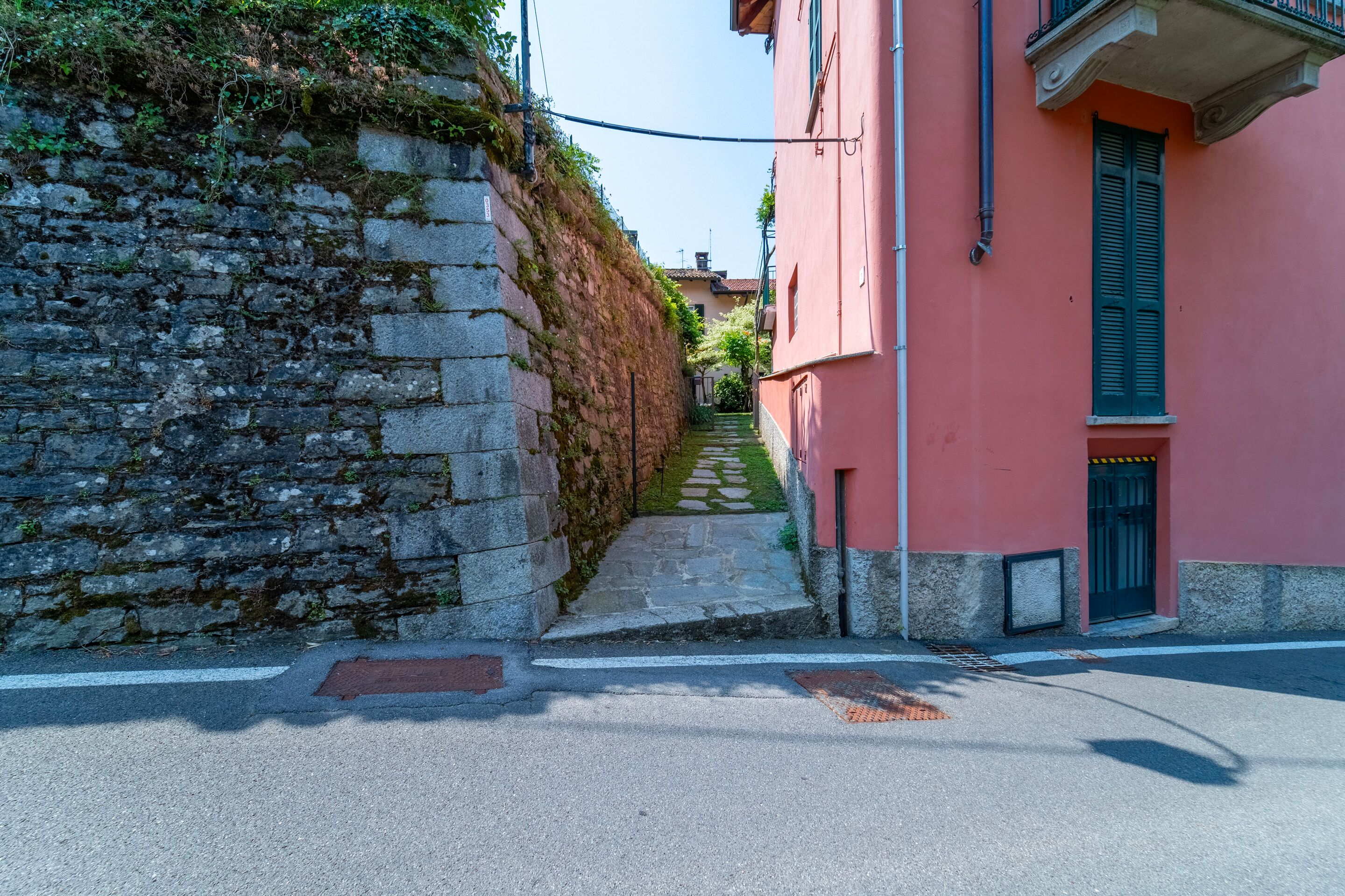 A narrow street with a stone wall on one side and a pink building with a balcony on the other.