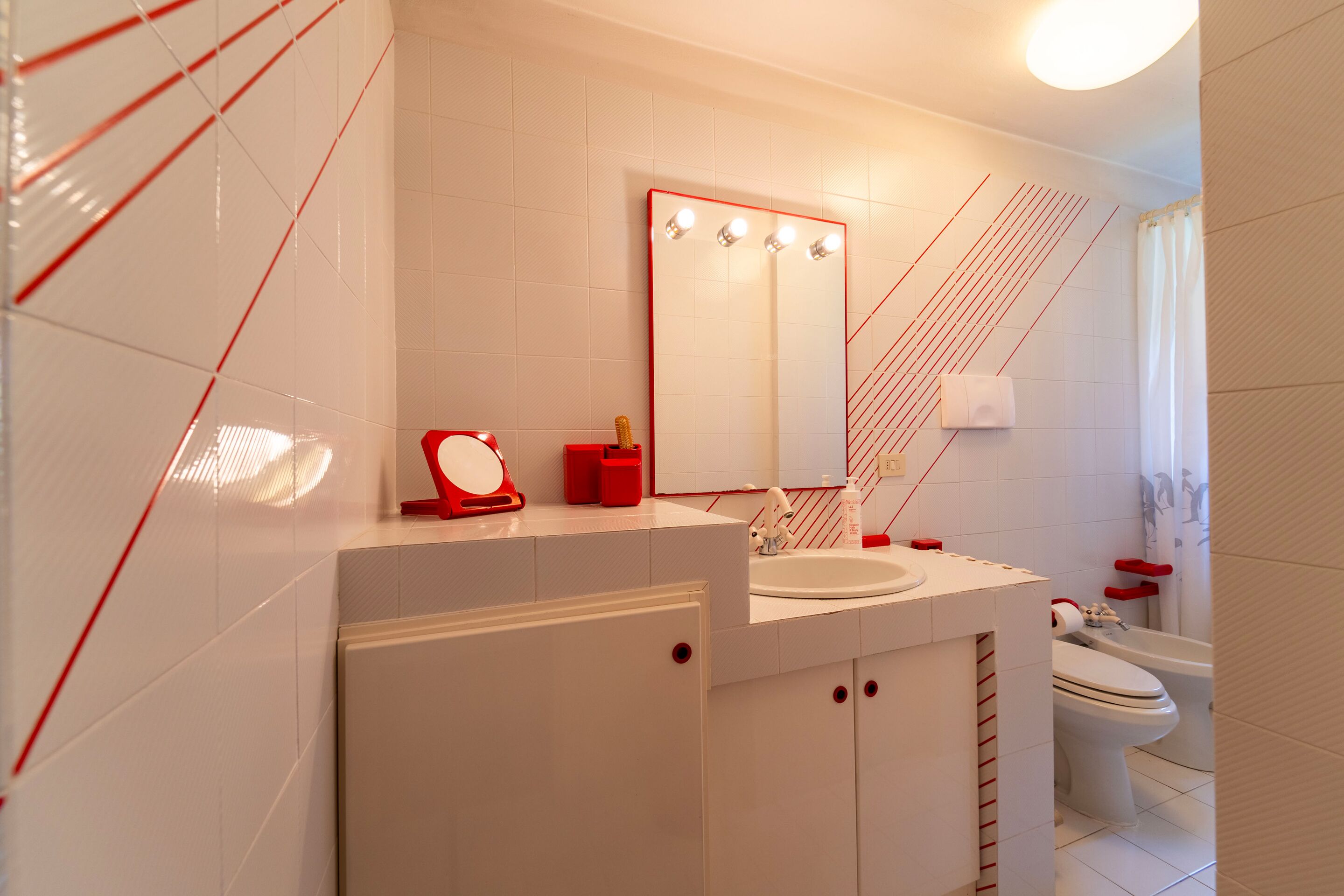 Un baño moderno con un lavamanos blanco, paredes con azulejos rojos, un espejo grande con luces y un tozalero rojo.