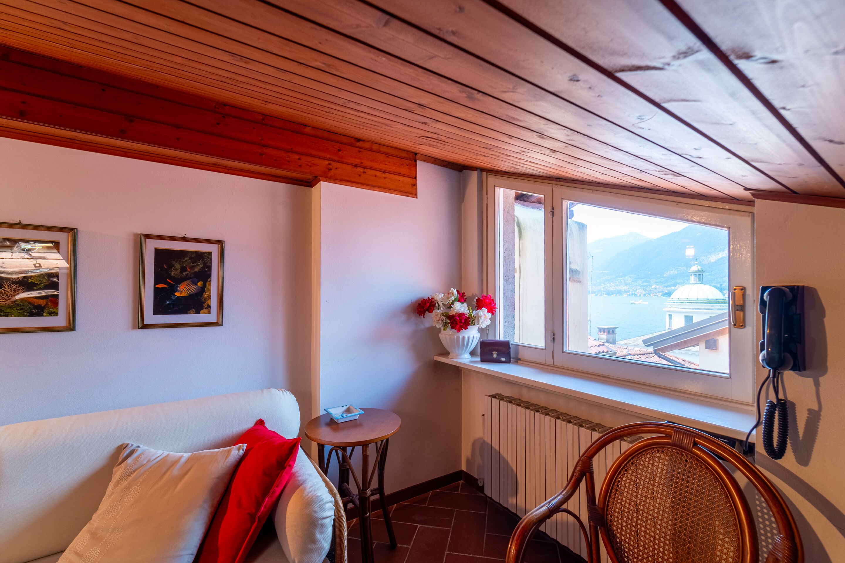 Een gezellige kamer met een houten plafond, een witte bank met rode kussens, een rieten stoel, een kleine tafel met een bloevas, en een raam met uitzicht op bergen en een gebouw.