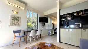 A modern living room with a wooden coffee table, two chairs, a dining table, and a kitchen area with cabinets and a TV.
