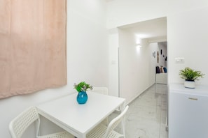 Une table blanche avec un vase bleu, des chaises blanches, un réfrigérateur blanc et un couloir avec une porte donnant sur une autre pièce.