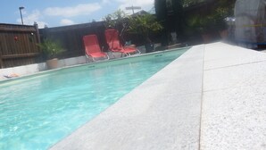 Una piscina con agua cristalina, rodeada por un patio de baldosas. Hay dos sillas reclinables rojas y algunas plantas en macetas.