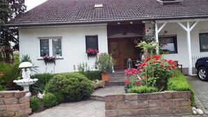 Una casa con techo de tejas, un porche delantero y un jardín con diversas plantas y flores.