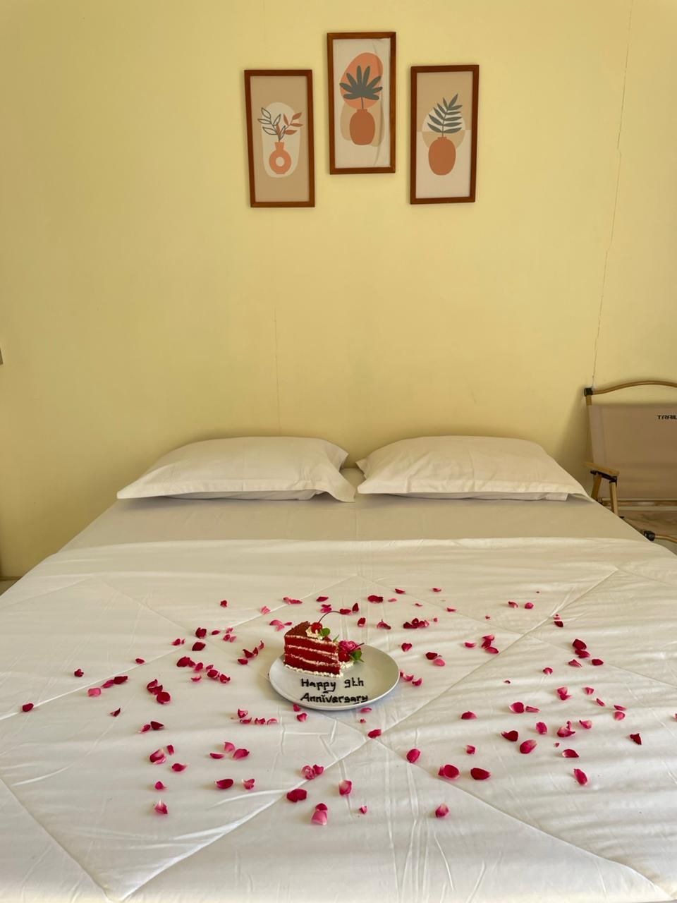 A bed with white linens, a cake on a plate with 'Happy 9th Anniversary' text, and rose petals scattered on the bed.