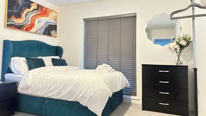 A bedroom with a teal bed, white bedding, a black dresser, a mirror, and a painting on the wall.