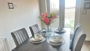 A dining table with a dark textured surface, surrounded by six grey upholstered chairs. A vase with pink flowers is placed on the table, and there are two white plates with folded napkins.