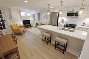 A modern kitchen with a central island, bar stools, a dining area with a wooden table and chairs, and a living area with a sofa and TV.
