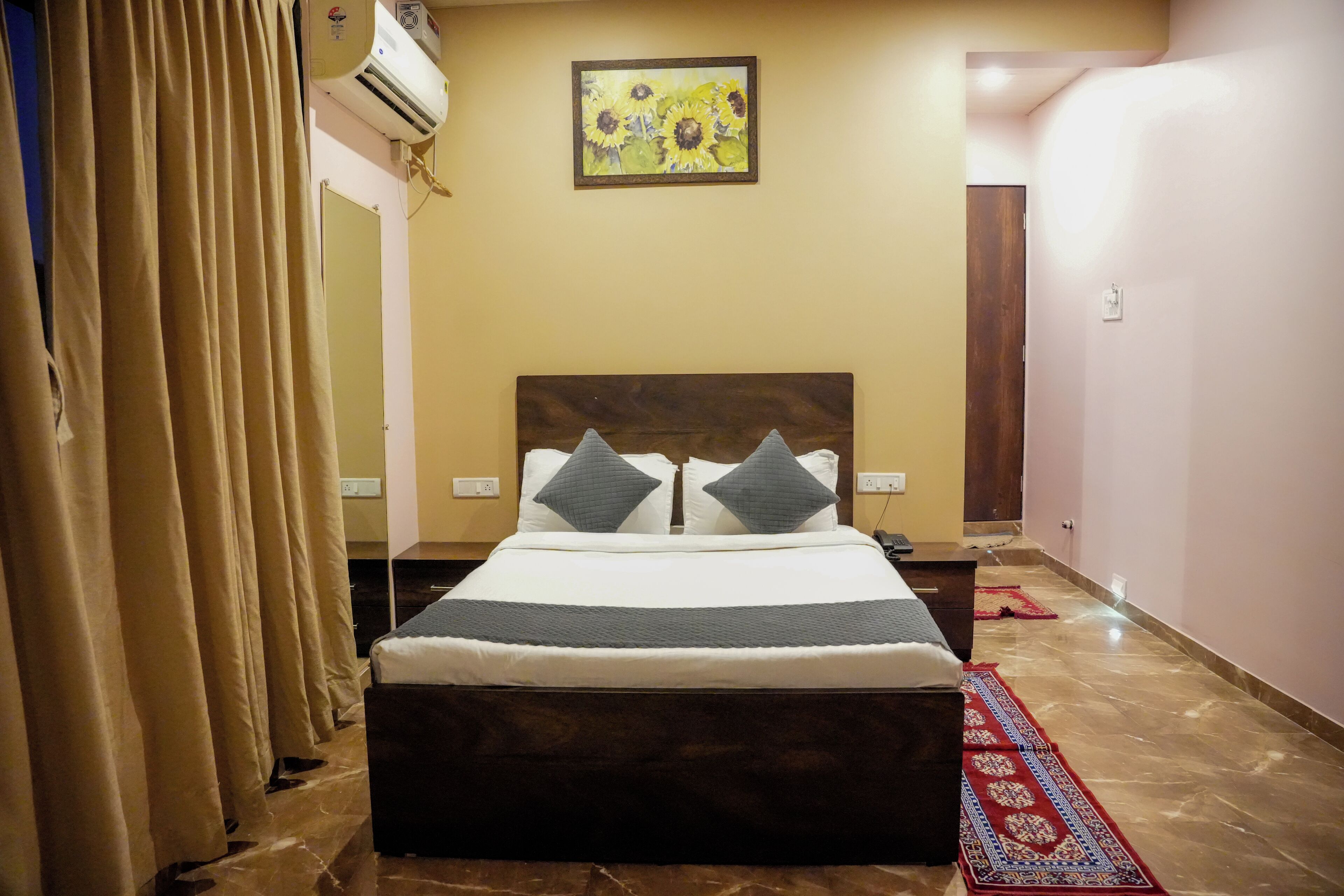 A hotel room with a bed, curtains, an air conditioner, a painting, and a red rug.