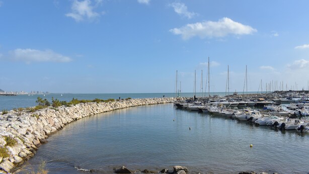 Una marina con barcos amarrados a lo largo de un muro de piedra curvo, agua tranquila y cielo despejado.