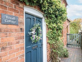 Uma cabana de tijolos com porta azul, uma placa com a inscrição 'Yoxall Cottage' e uma guirlanda na porta.