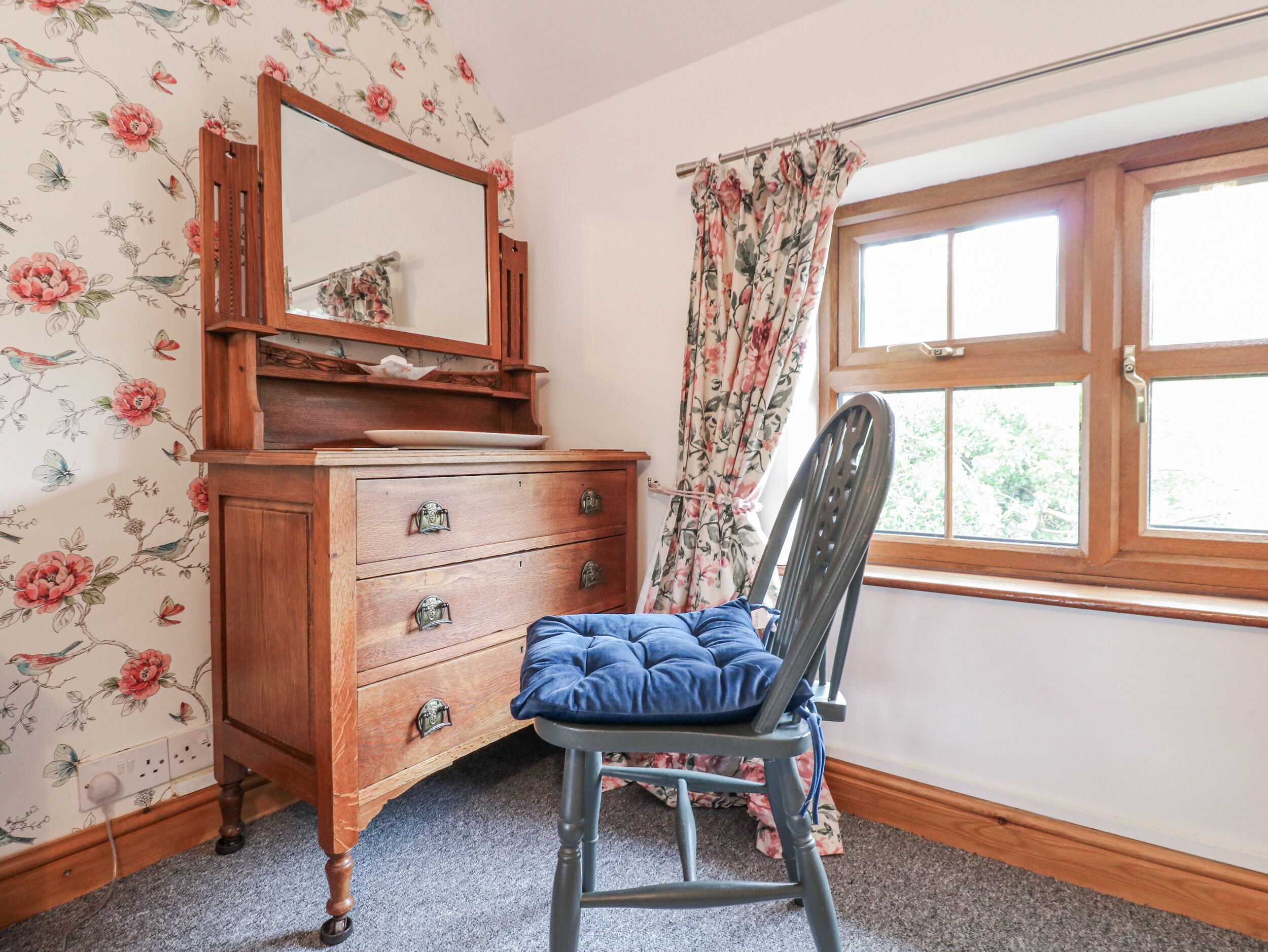A room with a wooden dresser, a mirror, a chair with a blue cushion, floral wallpaper, and a window with curtains.
