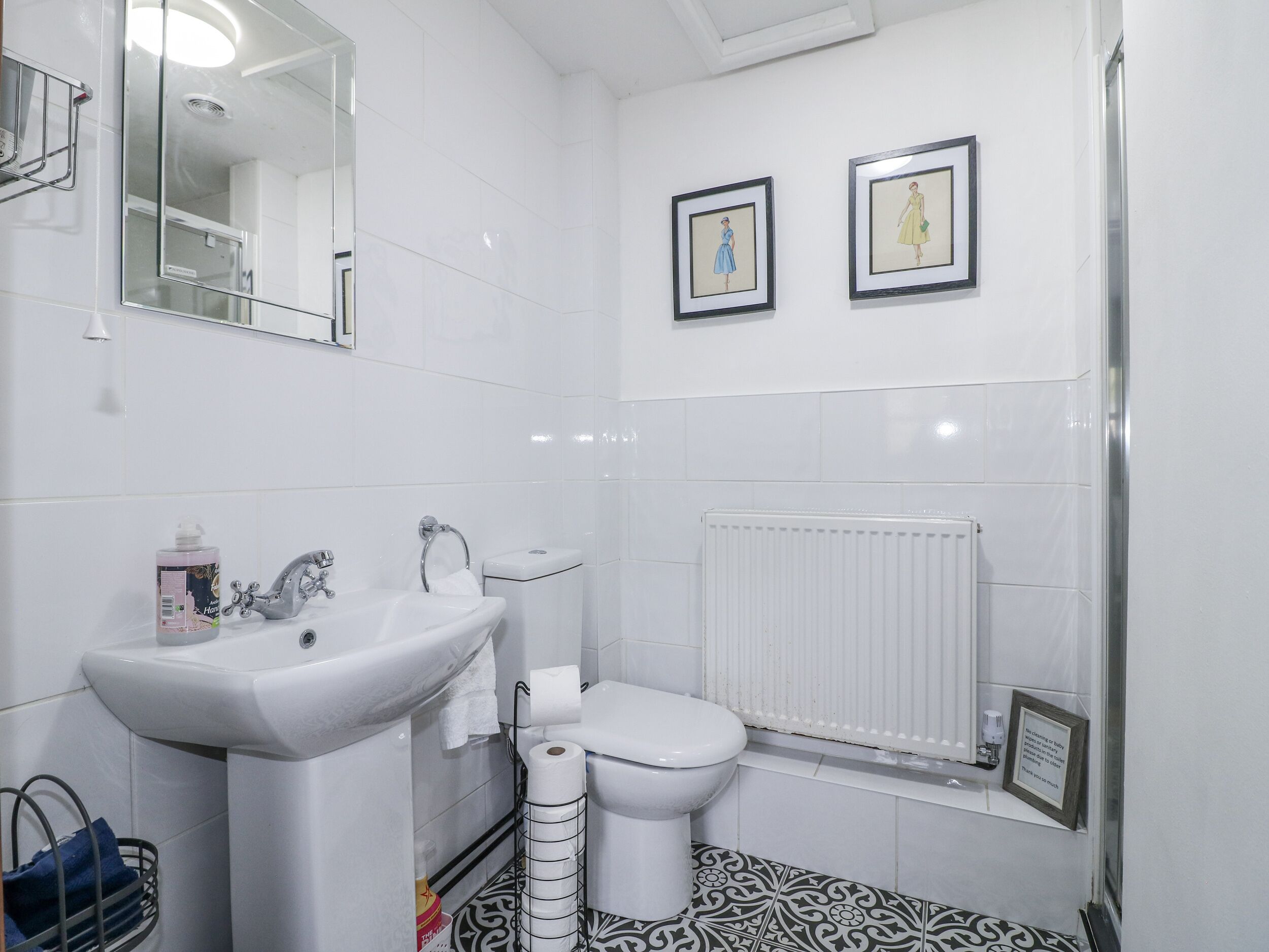 Un bany modern amb un lavabo, vàter i dutxa de color blanc. Hi ha dos quadres amb marc a la paret i un radiador sota el vàter.