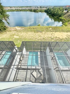 Blick auf einen Poolbereich mit klarem Wasser, umgeben von einer Rasenfläche und einem Gewässer im Hintergrund.