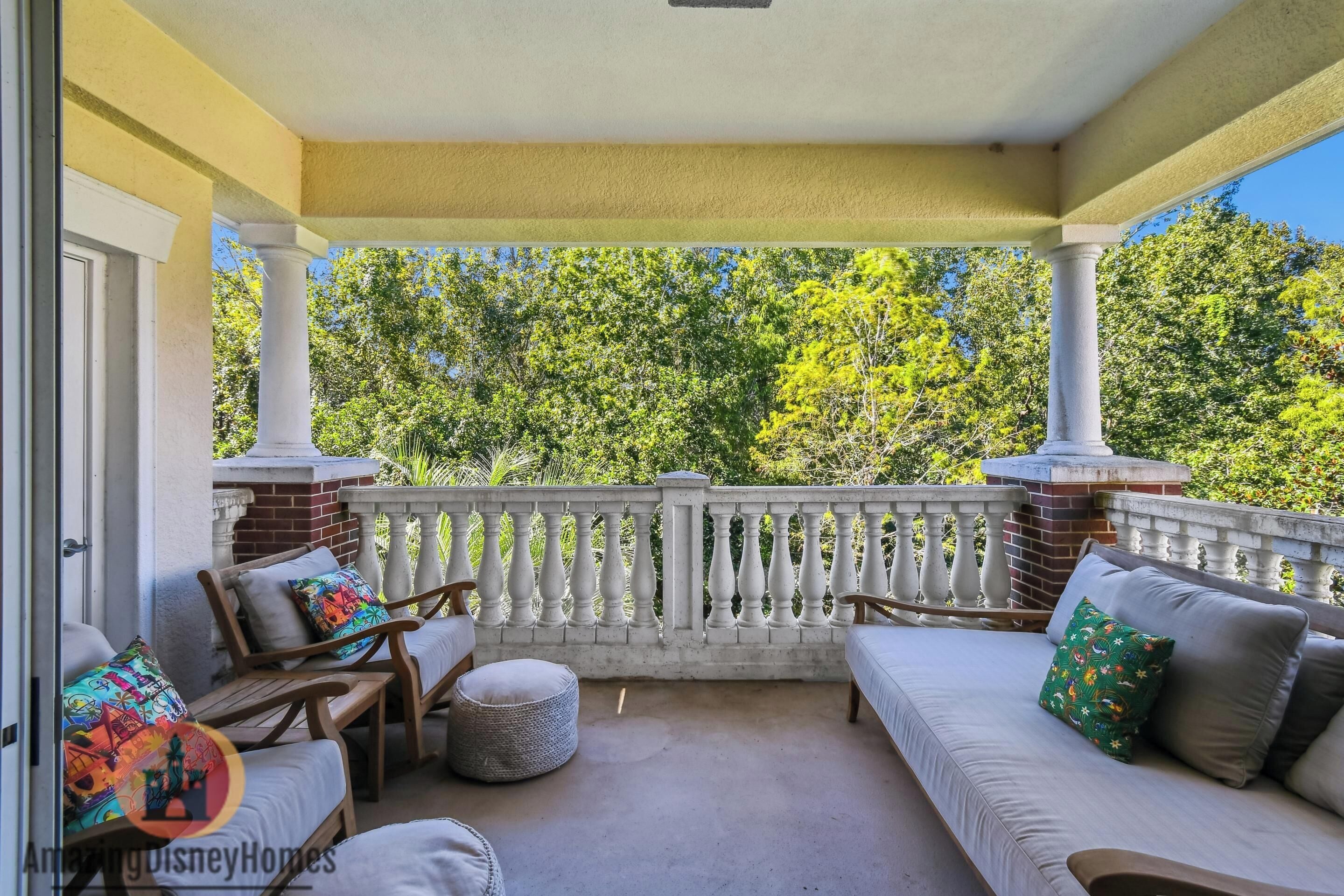 Une véranda couverte avec une balustrade blanche, deux chaises en bois, un canapé et un petit pouf rond.