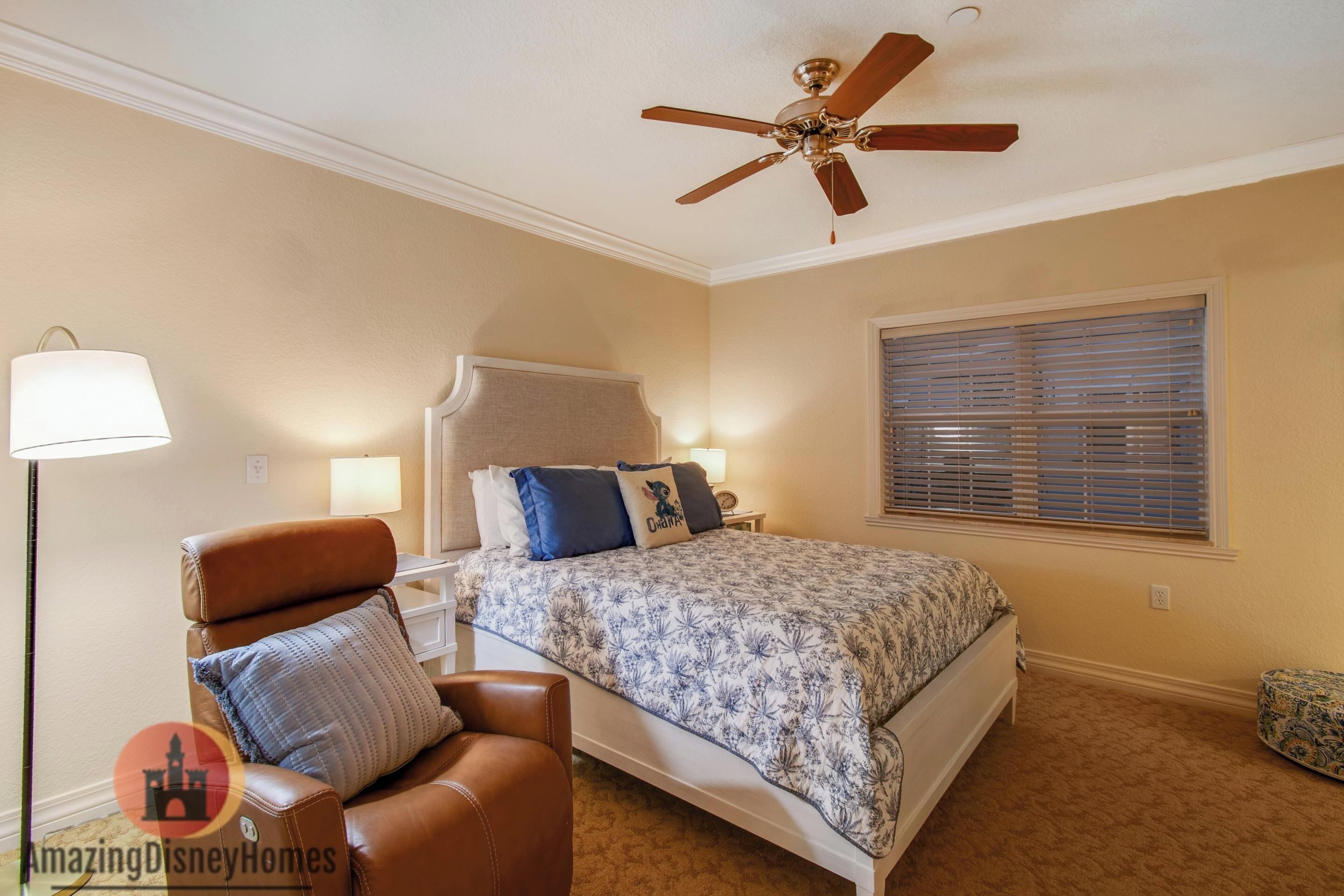 A bedroom with a bed, a ceiling fan, a nightstand, a lamp, a chair, and a window with blinds.
