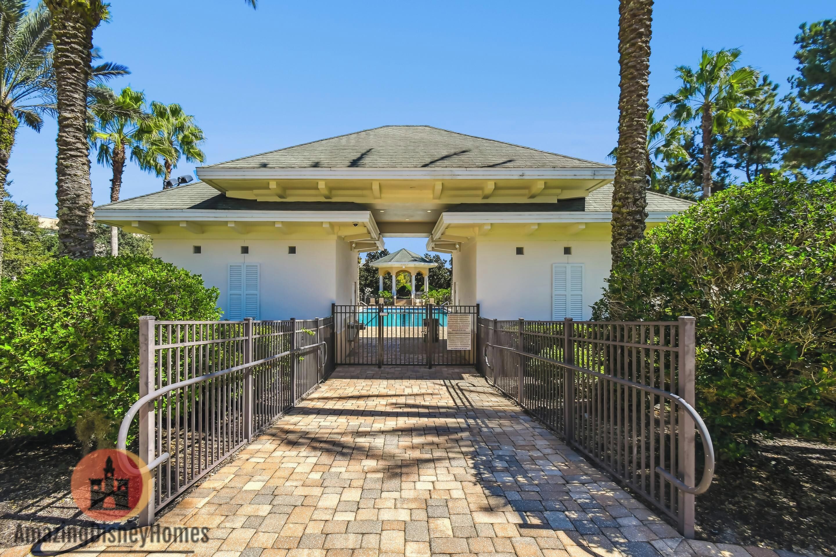 A pathway leading to a building with a pool, surrounded by palm trees and a gated entrance.
