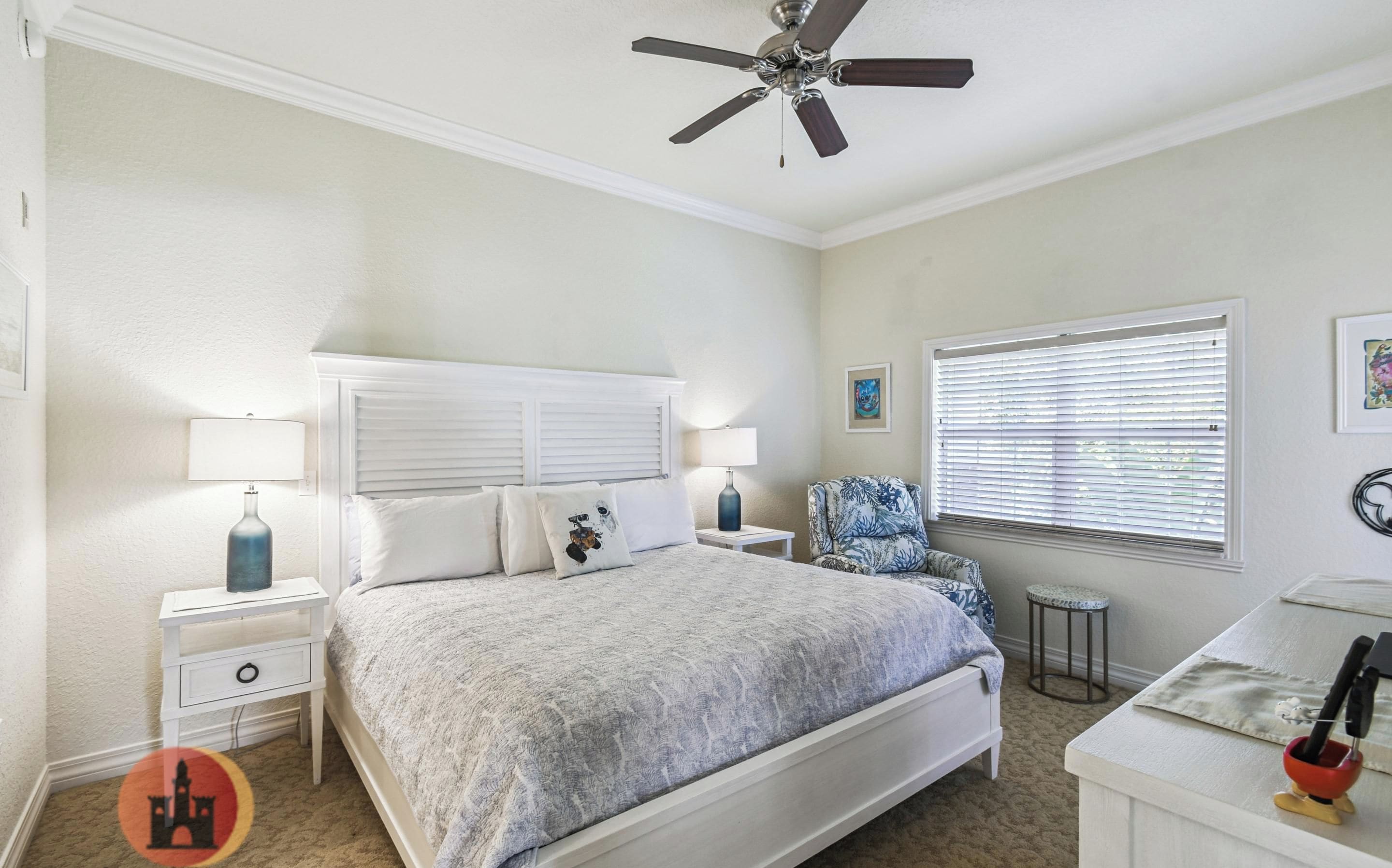 A bedroom with a bed, nightstands, a chair, a desk, a ceiling fan, and a window with blinds.