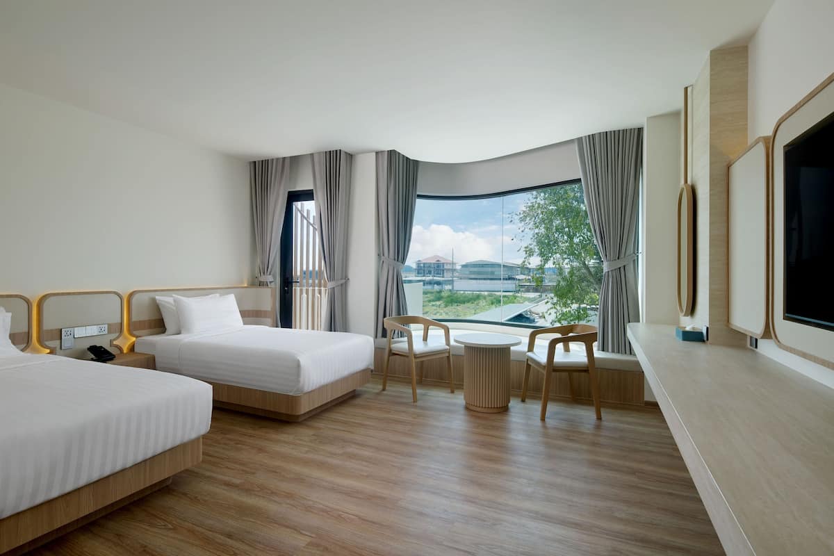 Ein Hotelzimmer mit zwei Betten, einem Esstisch, Stühlen, einem Fernseher und einem großen Fenster mit Blick auf Gebäude und Bäume.