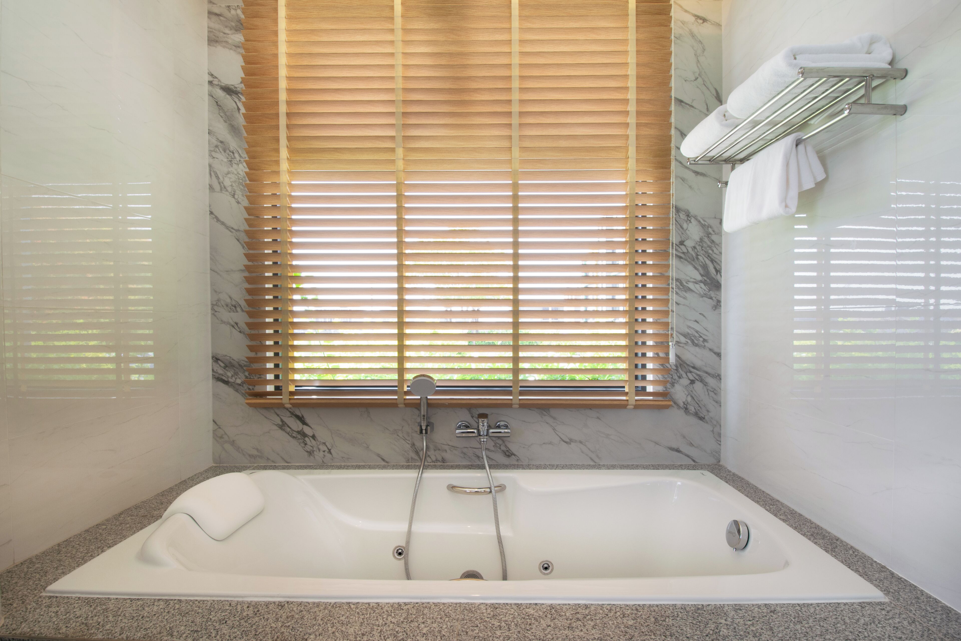 A modern bathroom with a marble wall, a large bathtub, wooden blinds, and a towel rack.