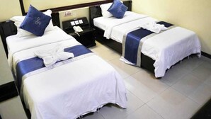 A hotel room with two single beds, each with a pillow and a blanket. There is a bedside table with a phone and a small lamp.