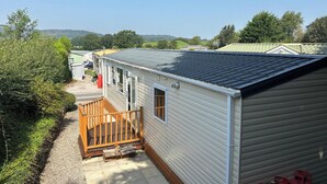 A mobile home with a wooden deck and a small porch, surrounded by greenery and other mobile homes in a rural setting. - 2 Bed Caravan-sleeps 6-pet Friendly (Carnforth)