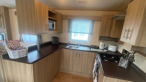 A compact kitchen with wooden cabinets, a sink, a stove, and a window with blinds. - 2 Bed Caravan-sleeps 6-pet Friendly (Carnforth)