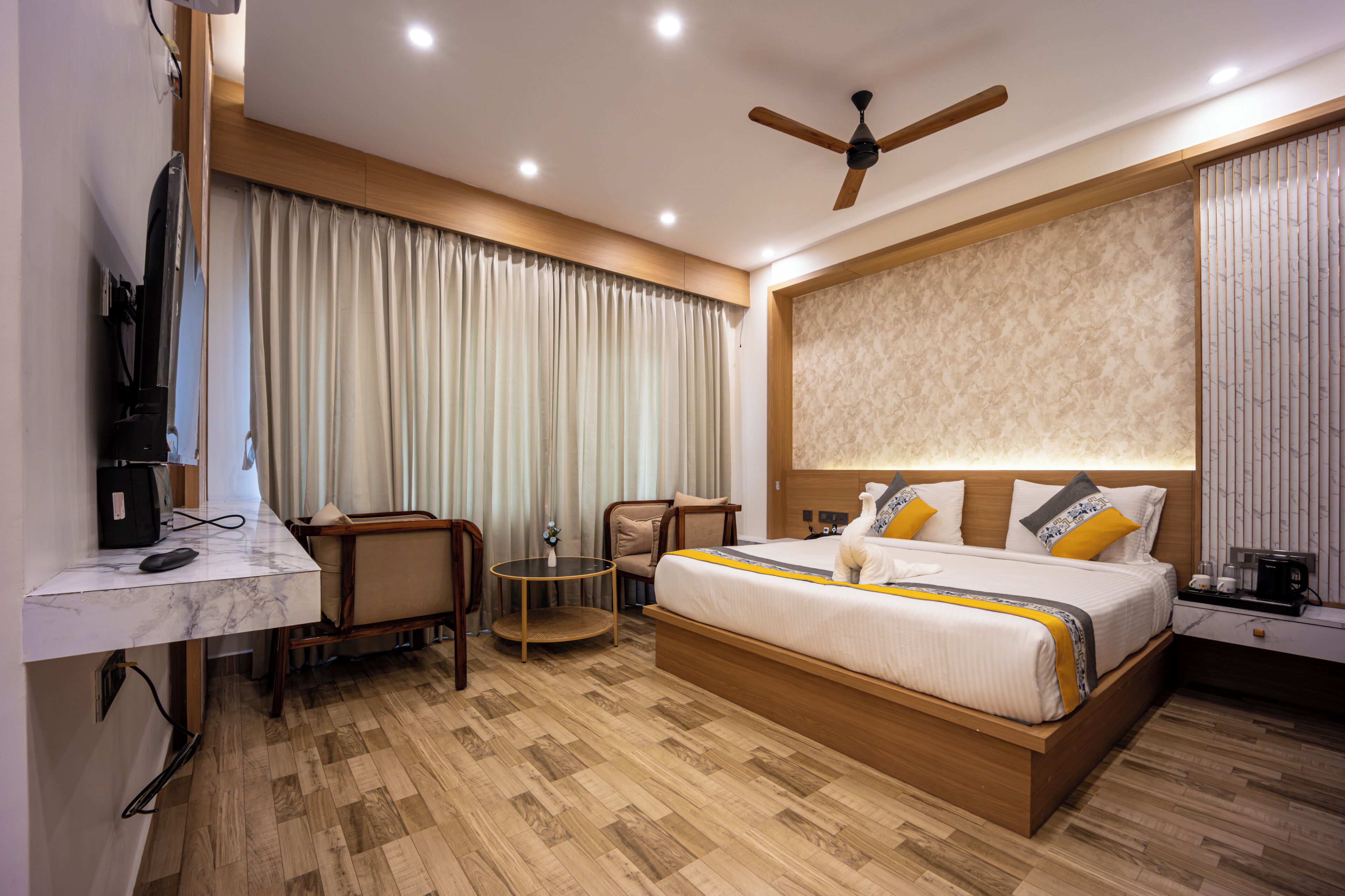 Une chambre d’hôtel moderne avec un lit en bois, un bureau avec un ordinateur, une chaise et un ventilateur de plafond.