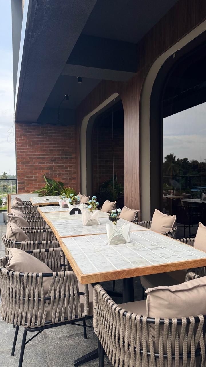 Un espacio de comedor al aire libre con una mesa larga y sillas de mimbre, frente a un muro de ladrillo con ventanas amplias.
