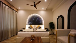 Une chambre d’hôtel avec un grand lit, une table basse en bois, un canapé, un ventilateur de plafond et une grande fenêtre ornée d’un motif de feuilles.