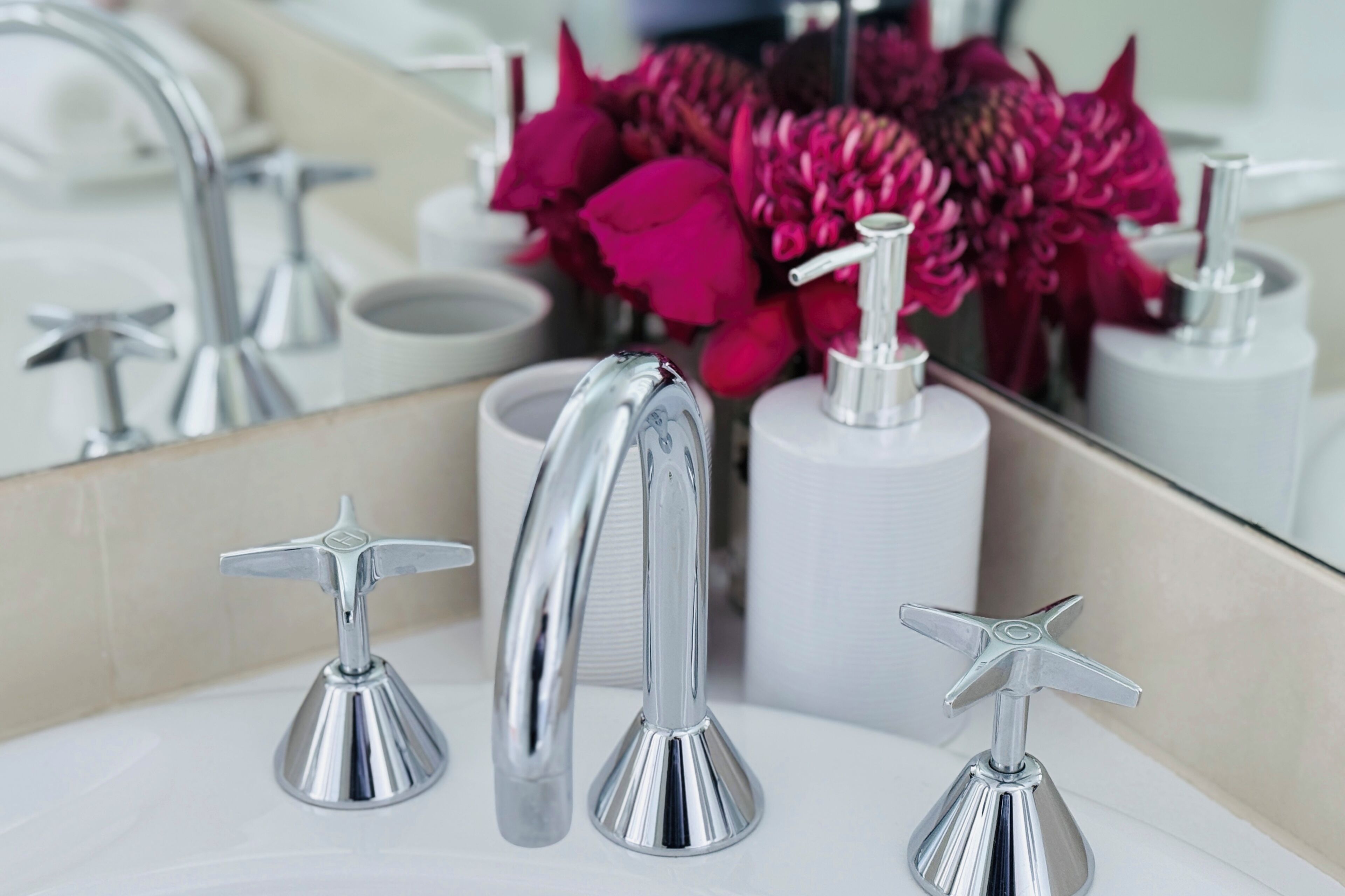 Un baño moderno con un espejo grande, tres grifos cromados y un jarrón de flores rojas.