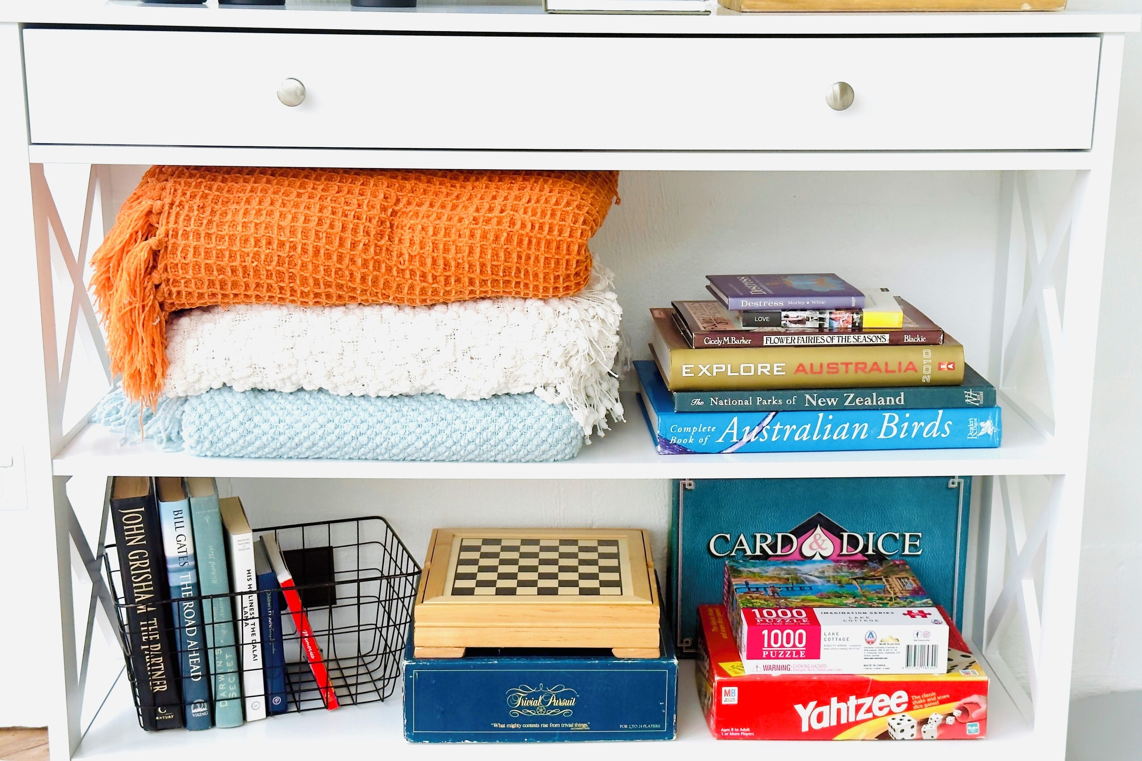 A white chest of drawers with a knitted blanket, books, a chessboard, and a card and dice game.