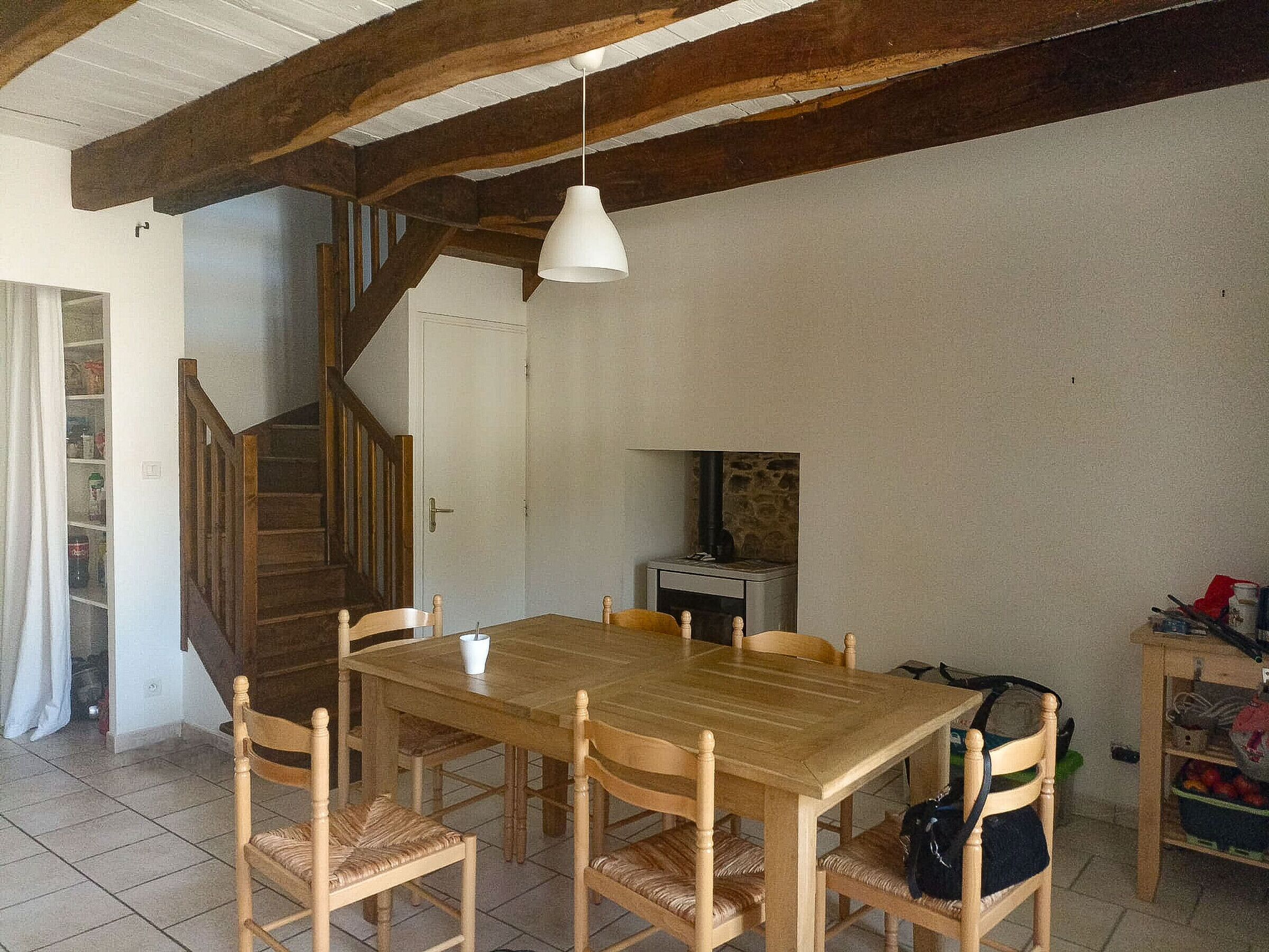 Een eethoek met een houten tafel en stoelen, een witte hanglamp, een trap en een klein keukentje met een fornuis.