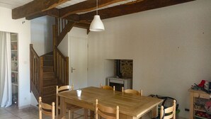 Un espace repas comprenant une table et des chaises en bois, un luminaire suspendu blanc, un escalier et une petite cuisine avec une cuisinière.