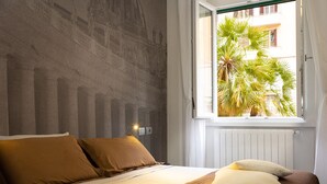 Camera da letto con un letto, cuscini, una finestra vista palme e una parete con motivo decorativo.