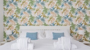 Habitación de hotel con cama, ropa blanca de cama, dos almohadas azules y un papel tapiz con estampado de hojas tropicales.